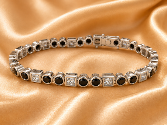 A silver tennis bracelet with alternating round black gemstones and square clear stones, displayed on a draped gold satin fabric background.