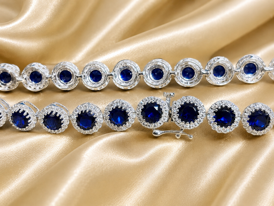 A close-up of a silver tennis bracelet featuring round deep blue gemstones surrounded by clear crystal halo settings, displayed on a draped champagne satin fabric.