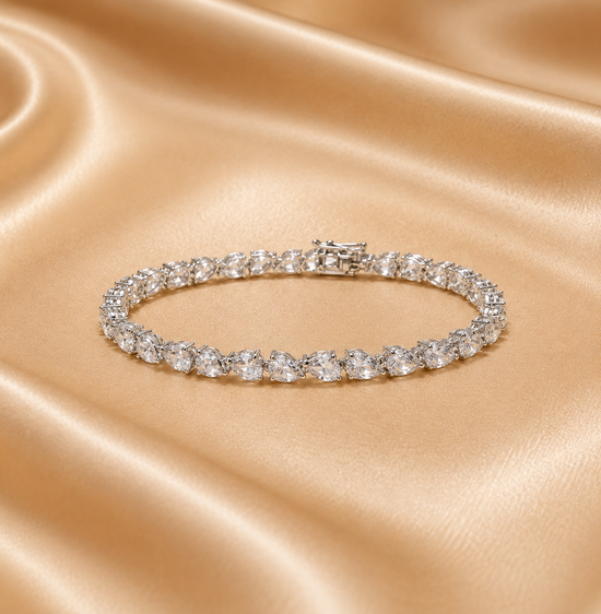 A tennis bracelet with sparkling clear stones set in a silver-toned metal, displayed on a draped champagne satin fabric background.