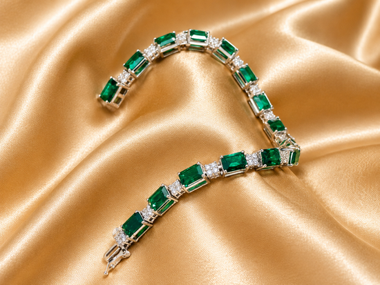 A silver tennis bracelet featuring emerald-cut green gemstones and small clear accent stones, arranged on a draped gold satin fabric background.