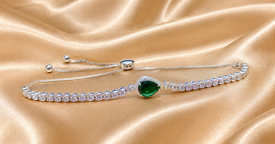 A silver-tone adjustable bracelet featuring a central heart-shaped green emerald gemstone surrounded by small clear stones, displayed on a draped champagne satin fabric.