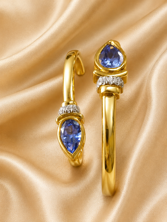 A pair of gold hoop earrings with pear-cut blue gemstones and small clear accent stones, displayed on a draped champagne satin fabric.