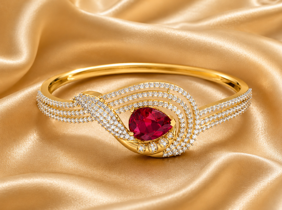 A gold bangle bracelet featuring a central pear-cut red ruby gemstone, surrounded by a double row of pavé-set clear cubic zirconia stones, displayed on a draped gold satin fabric.