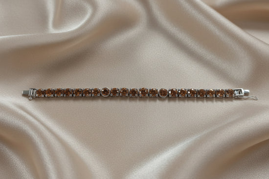 A silver tennis bracelet with round brown gemstones, displayed horizontally on a draped beige satin fabric background.