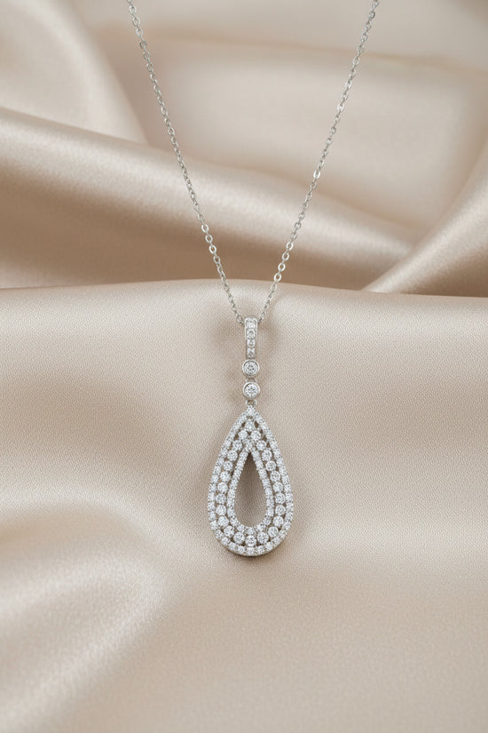 A silver teardrop pendant necklace paved with small clear stones, suspended from a delicate silver chain on a white background with a soft reflection below.