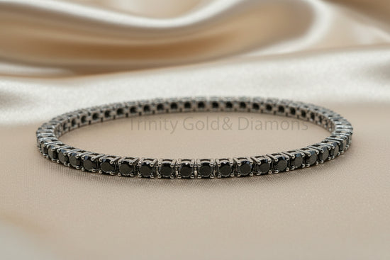 A single tennis bracelet with round black gemstones in a silver-toned setting, displayed on a beige satin fabric background. A faint &