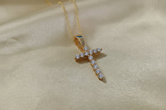A gold cross pendant adorned with small clear stones, hanging from a delicate gold chain on a beige satin fabric background.
