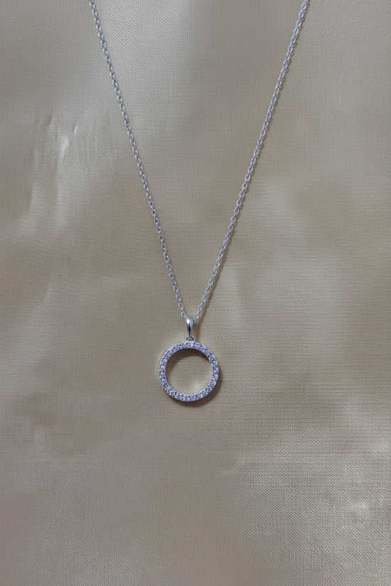 A silver chain necklace with a circular pendant paved with small clear stones, displayed on a beige satin fabric background.