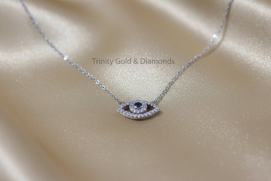 A silver-toned evil eye necklace with a dark blue center stone and clear pavé accents, displayed on a shimmering champagne satin fabric. The brand name &