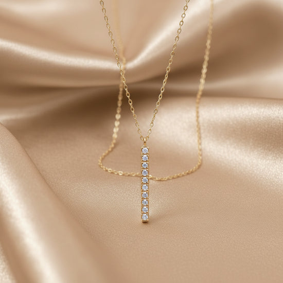 A delicate gold necklace with a vertical bar pendant set with small clear stones, displayed on draped champagne satin fabric.