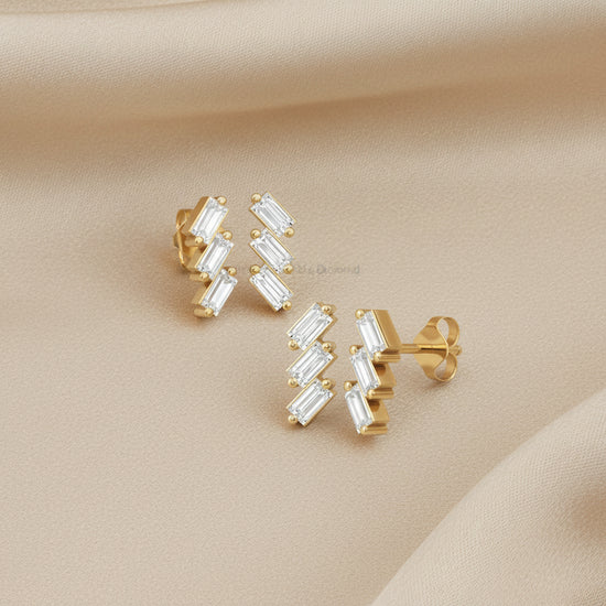 A pair of gold stud earrings with a zigzag arrangement of clear baguette-cut gemstones, displayed on a draped beige fabric background.