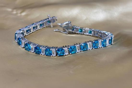 A silver tennis bracelet featuring rectangular blue gemstones, displayed on a draped beige satin fabric background.