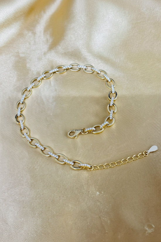 A gold chain bracelet with small white inlaid accents, displayed on a draped beige satin fabric background.