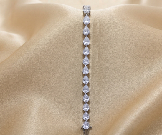 A silver tennis bracelet featuring a vertical line of pear-cut clear gemstones, displayed on draped champagne-colored satin fabric.