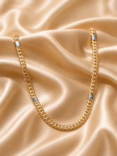 A gold-toned Cuban link chain necklace with silver-toned rectangular end caps, draped on a shiny champagne satin fabric background.