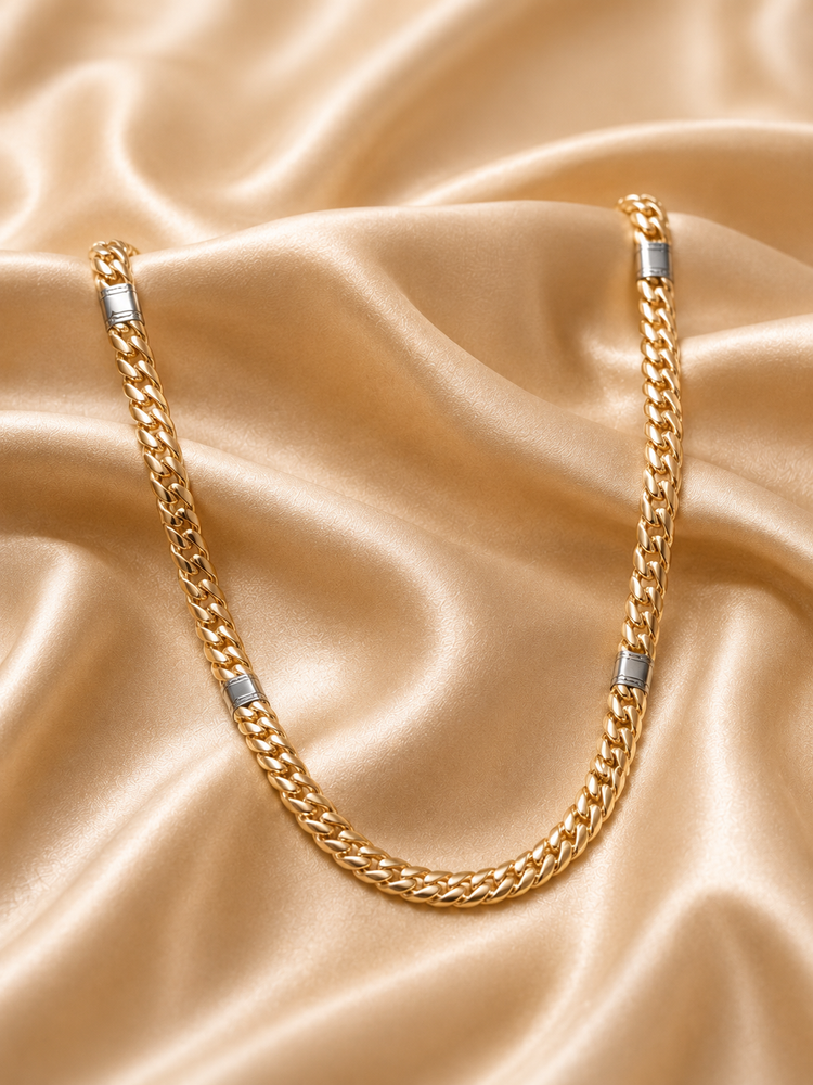A gold-toned Cuban link chain necklace with silver-toned rectangular end caps, draped on a shiny champagne satin fabric background.