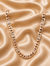 A chunky gold-toned Cuban link chain necklace with rectangular links, displayed on a draped champagne satin fabric background.