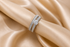 A silver-toned double band ring with a round-cut center stone and pavé-set diamonds, displayed on a finger against a draped champagne silk fabric background.