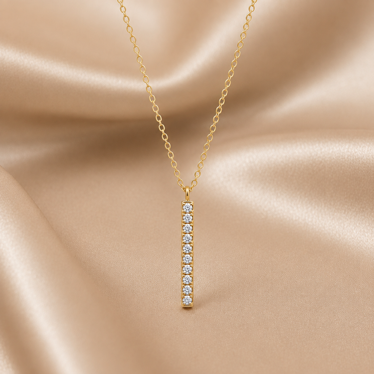 A gold chain necklace with a slim vertical bar pendant set with small clear stones, displayed against a draped champagne satin fabric background.