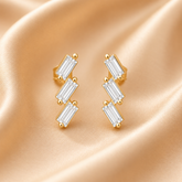 Gold Earrings with Three Baguette-Cut Clear Stones in a Zigzag Design on a Champagne Silk Background
