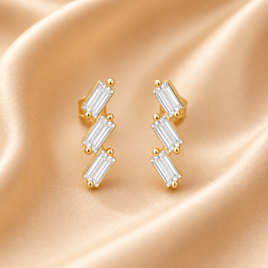 Gold Earrings with Three Baguette-Cut Clear Stones in a Zigzag Design on a Champagne Silk Background

