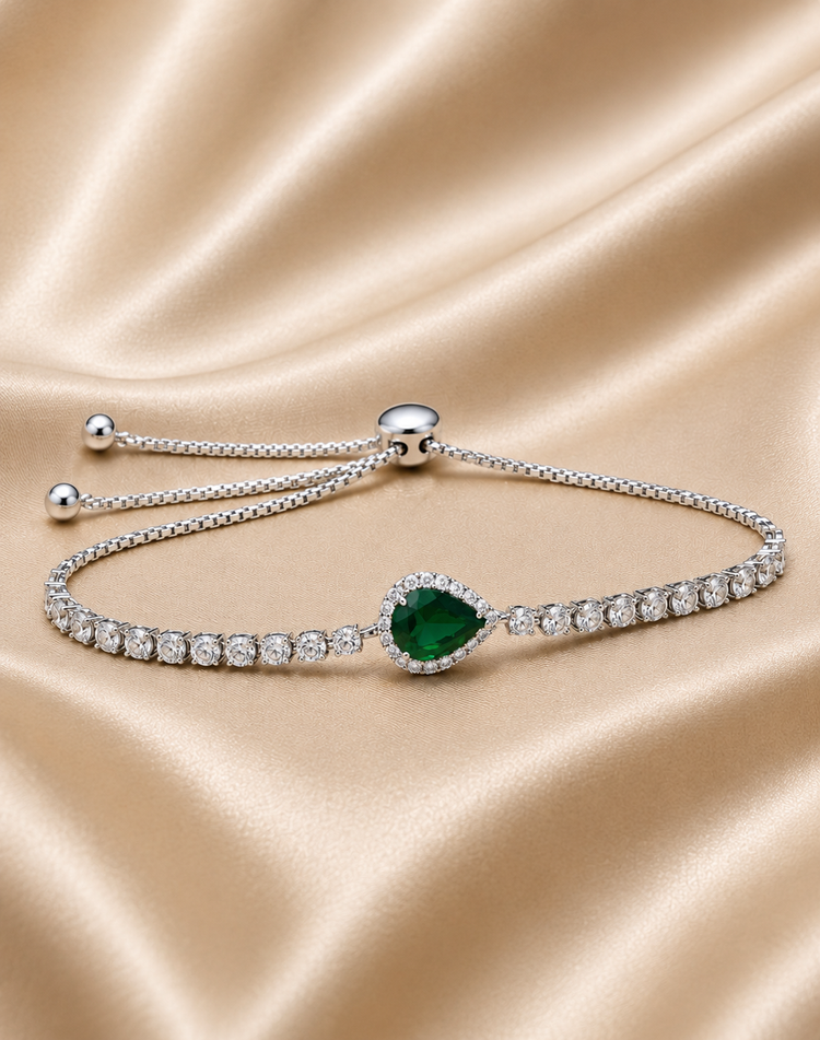 A silver-tone adjustable bracelet featuring a central heart-shaped green emerald gemstone surrounded by small clear stones, displayed on a draped champagne satin fabric.