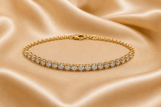 A gold tennis bracelet with a row of clear round-cut gemstones, displayed on a draped champagne-colored satin fabric.