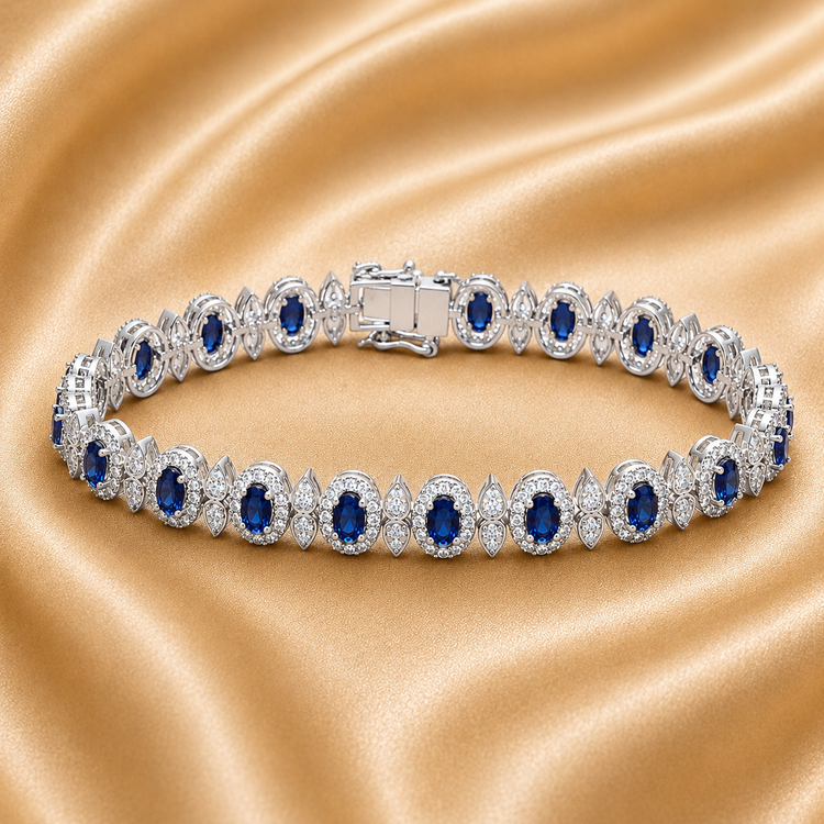 A silver tennis bracelet with oval blue gemstones and clear stones, displayed on a draped champagne satin fabric.