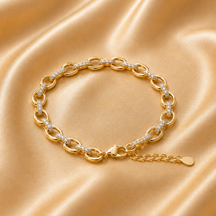 A gold chain bracelet with small white inlaid accents, displayed on a draped beige satin fabric background.