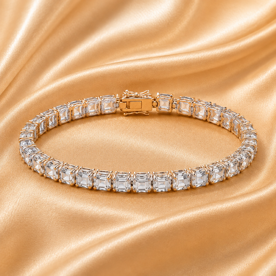 A curved tennis bracelet with baguette-cut clear stones in a silver-toned setting, displayed on draped champagne satin fabric.