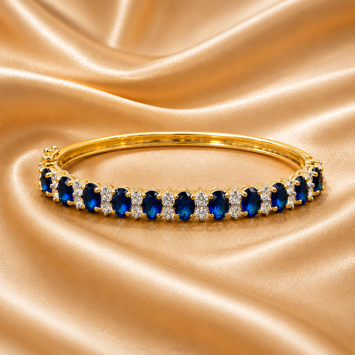 A gold bangle bracelet featuring a row of oval-cut deep blue gemstones and small clear accent stones, displayed on a draped beige satin fabric background.