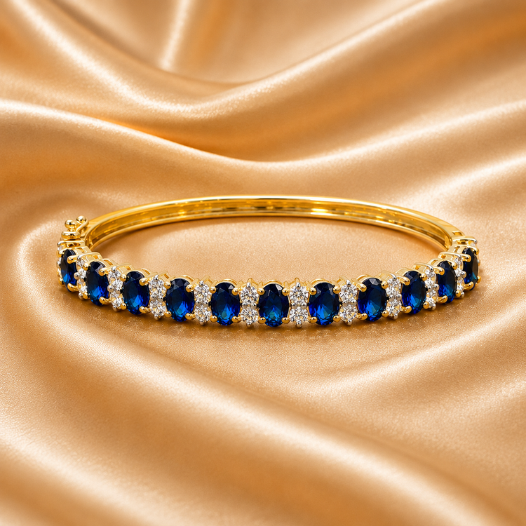 A gold bangle bracelet featuring a row of oval-cut deep blue gemstones and small clear accent stones, displayed on a draped beige satin fabric background.