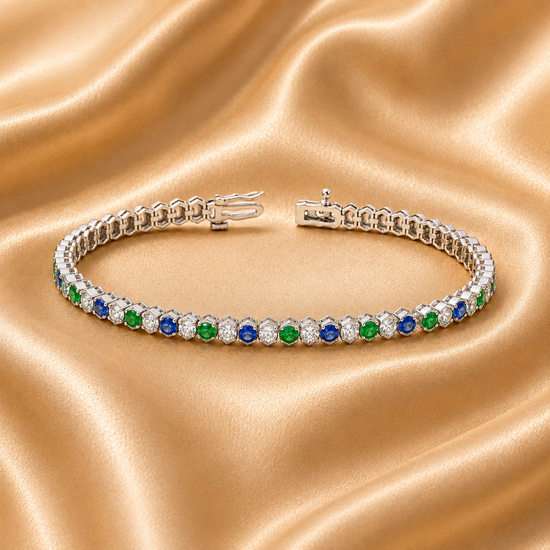 A silver tennis bracelet with alternating round blue and green gemstones and clear accent stones, displayed on a draped beige satin fabric background.