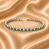 A silver tennis bracelet with alternating round blue and green gemstones and clear accent stones, displayed on a draped beige satin fabric background.