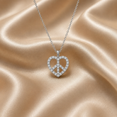 A silver peace sign pendant necklace encrusted with small clear stones, held in the palm of a hand against a draped champagne satin fabric background.