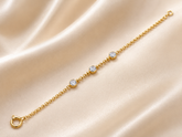 A delicate yellow gold chain bracelet with three round-cut clear gemstones, displayed diagonally on a draped beige satin fabric background.