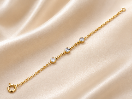 A delicate yellow gold chain bracelet with three round-cut clear gemstones, displayed diagonally on a draped beige satin fabric background.