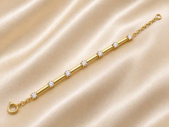 A gold bracelet with five round-cut clear gemstones spaced along a polished tube, displayed on a draped beige satin fabric background.