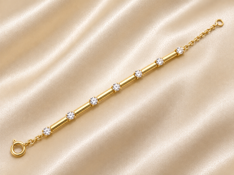 A gold bracelet with five round-cut clear gemstones spaced along a polished tube, displayed on a draped beige satin fabric background.