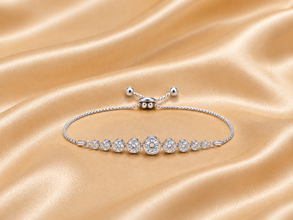 A silver-toned bracelet with a row of round clear gemstones in bezel settings, displayed on a draped champagne satin fabric background.