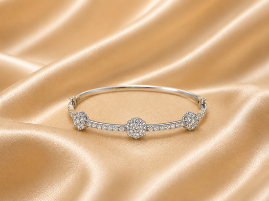 A silver-toned bangle bracelet with three floral diamond clusters and pavé accents, displayed on draped champagne satin fabric.