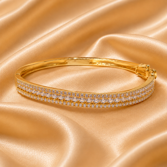 A gold bangle bracelet with two rows of pavé-set clear stones, displayed on a draped champagne satin fabric background.