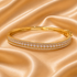 A gold bangle bracelet with two rows of pavé-set clear stones, displayed on a draped champagne satin fabric background.