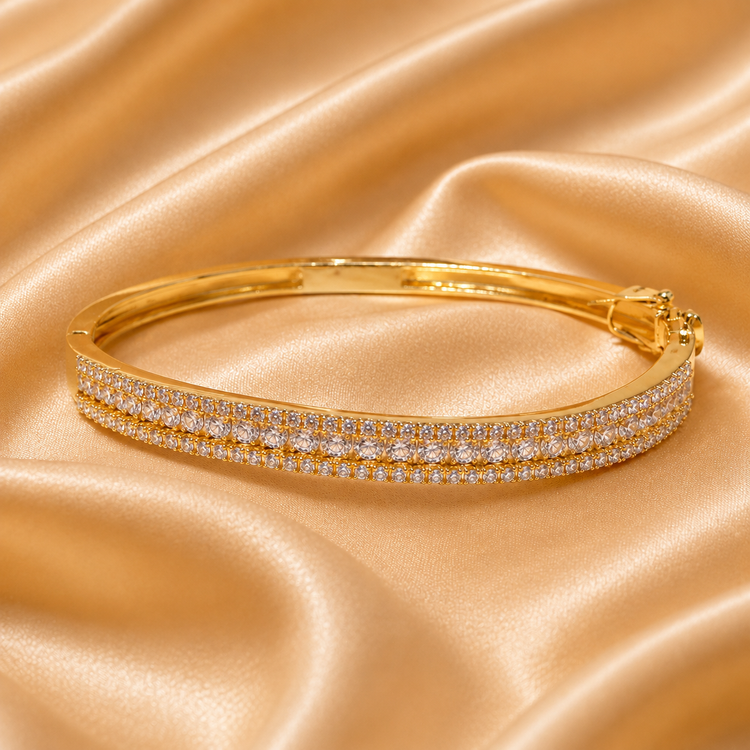 A gold bangle bracelet with two rows of pavé-set clear stones, displayed on a draped champagne satin fabric background.