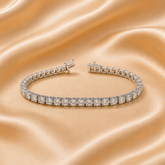 A tennis bracelet with clear gemstones in a silver-toned setting, displayed on a draped champagne satin fabric background.