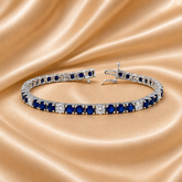 A tennis bracelet with round blue sapphires and clear accent stones in a silver-toned setting, displayed on a draped champagne satin fabric background.