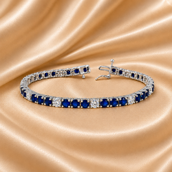 A tennis bracelet with round blue sapphires and clear accent stones in a silver-toned setting, displayed on a draped champagne satin fabric background.
