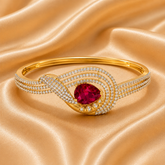 A gold bangle bracelet featuring a central oval-cut deep pinkish-red gemstone, surrounded by a swirling design of pavé-set clear crystals and a polished gold band, displayed on a draped champagne satin fabric.