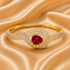A gold bangle bracelet featuring a central oval-cut deep pinkish-red gemstone, surrounded by a swirling design of pavé-set clear crystals and a polished gold band, displayed on a draped champagne satin fabric.