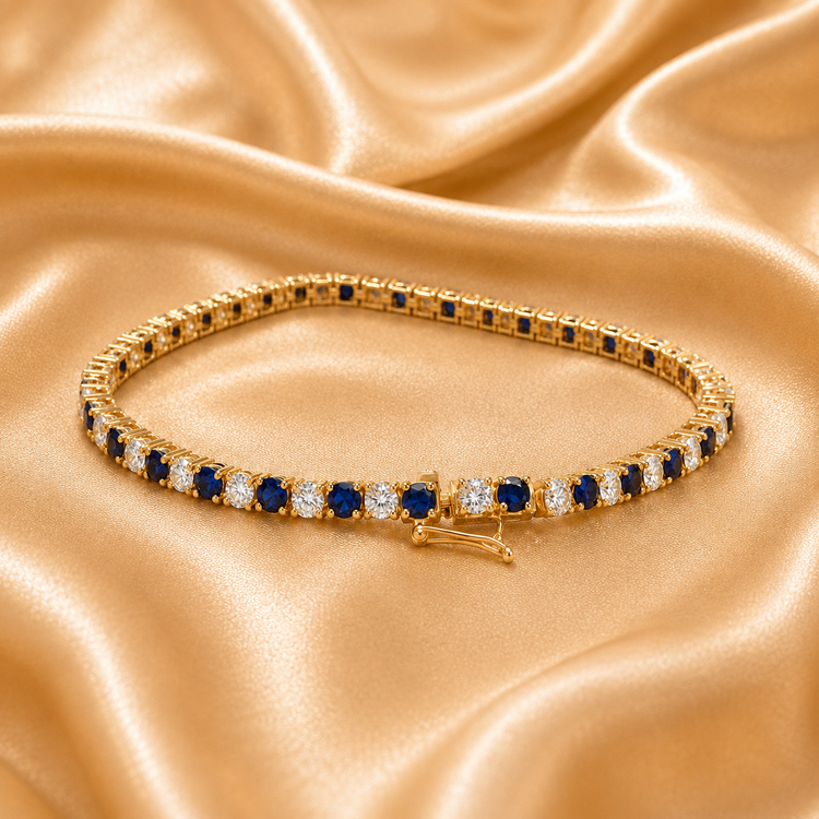 A gold tennis bracelet adorned with round blue sapphires and clear cubic zirconia stones, displayed on a draped champagne satin fabric.